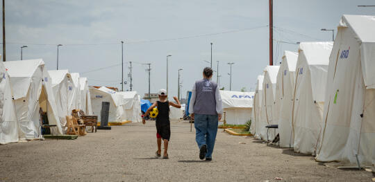 CH11629107 Heriberto (7) walks alongside a Save the Children staff among a row of tents