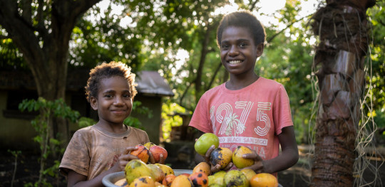 CH11271777 Madleen, 11, and Agnes, 7, holding mangoes outside their home on an island in Shefa Province, Vanuatu. (1)